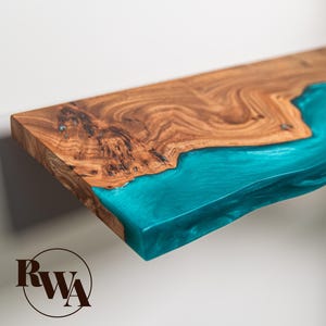 May include: A rectangular wooden shelf featuring a natural live edge and a smooth, vibrant turquoise resin inlay that flows across the surface. The wood displays a prominent brown grain pattern, and a circular logo with the letters RWA is visible.