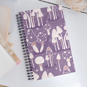 Puede incluir: Cuaderno de espiral con una cubierta morada adornada con un patrón repetido de setas blancas y beige. El cuaderno tiene una encuadernación en espiral negra. El diseño presenta varias formas y tamaños de setas.