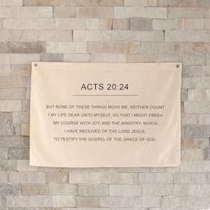 Puede incluir: Un tapiz de tela beige con el texto "ACTOS 20:24" y un versículo bíblico impreso en negro. El tapiz está montado en una pared de piedra gris claro.