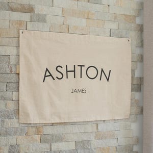 Puede incluir: Una pancarta de lona beige con el nombre "ASHTON" en fuente grande, negra y sin serifa, y "JAMES" debajo en una fuente más pequeña. La pancarta está colgada en una pared de piedra clara texturizada.
