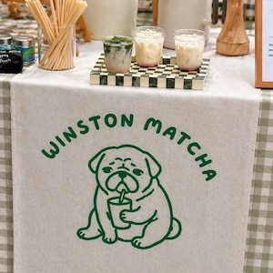 May include: A cream-coloured table runner featuring the words "WINSTON MATCHA" and a green pug illustration. The table is covered with a green and white checked tablecloth. Several drinks are displayed on the table.