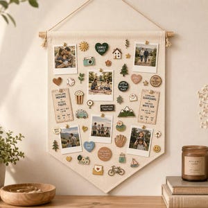 May include: A cream-colored fabric wall hanging banner featuring a wooden dowel and string, decorated with various enamel pins and five square photographs. The pins include shapes like a camera, house, mountains, bicycle, and hearts with text.