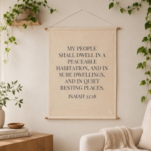 May include: A beige wall hanging with the text "MY PEOPLE SHALL DWELL IN A PEACEABLE HABITATION, AND IN SURE DWELLINGS, AND IN QUIET RESTING PLACES. ISAIAH 32:18." The wall hanging is suspended by wooden dowels. The image also shows a white armchair and plants.