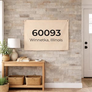 Op de afbeelding: Een beige wandkleed met de tekst "60093 Winnetka, Illinois" in zwart. Het wandkleed hangt aan een stenen muur. Een houten tafel met twee rieten manden staat eronder.