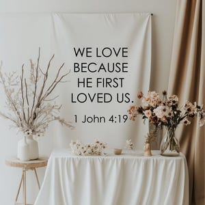 May include: A white banner with the text "WE LOVE BECAUSE HE FIRST LOVED US. 1 John 4:19" is displayed. The banner is hanging above a table draped with a white cloth. Floral arrangements and decorative items are on the table and a small side table.
