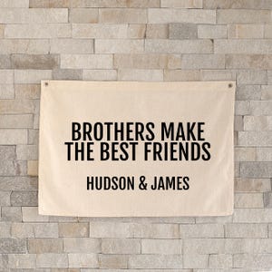 May include: A beige canvas banner with the text "BROTHERS MAKE THE BEST FRIENDS" in black, and the names "HUDSON & JAMES" below. The banner is mounted on a light-coloured brick wall.