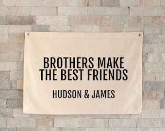 May include: A beige canvas banner with the text "BROTHERS MAKE THE BEST FRIENDS" in black, along with the names "HUDSON & JAMES" below. The banner is mounted on a light-colored brick wall.