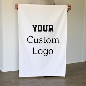 Puede incluir: Una pancarta blanca con las palabras "YOUR Custom Logo" impresas en negro. La pancarta está sujeta por una persona. El fondo es una pared blanca lisa.
