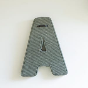 May include: A large, padded letter "A" in a muted green fabric with a corded texture. The letter has a cutout triangle and a dark leather strap for hanging. The edges are neatly finished with a contrasting trim.