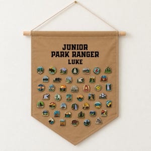 May include: A tan canvas wall banner featuring the black text JUNIOR PARK RANGER LUKE at the top. The banner is decorated with numerous small, colourful enamel pins depicting various national park landscapes, wildlife, and natural landmarks.