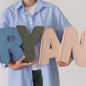 May include: Four large, plush letters spelling "RYAN" in shades of blue denim, olive green, and light pink. The letters are being held by a person wearing a blue and white striped shirt.