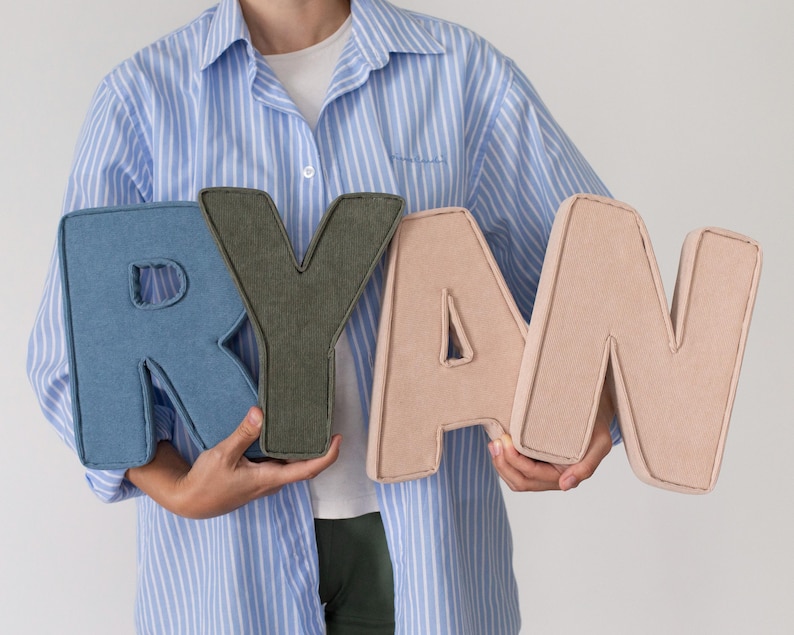 May include: Four large, plush letters spelling "RYAN" in shades of blue denim, olive green, and light pink. The letters are being held by a person wearing a blue and white striped shirt.