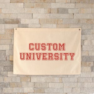 Puede incluir: Una pancarta beige con las palabras "CUSTOM UNIVERSITY" en letras rojas. La pancarta está colgada en una pared de ladrillos de color claro. Tiene dos ojales metálicos en la parte superior.