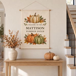 May include: A rectangular fabric wall hanging featuring a printed illustration of three pumpkins surrounded by autumn leaves and berries. The text reads "WELCOME TO THE MATTISON PUMPKIN PATCH" in dark brown lettering. The banner is suspended by a wooden rod.