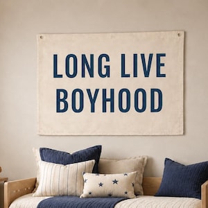 May include: A beige canvas banner with the words "LONG LIVE BOYHOOD" in navy blue lettering hangs above a bed with navy blue and patterned pillows. The bed has a navy blue blanket and a wooden frame.