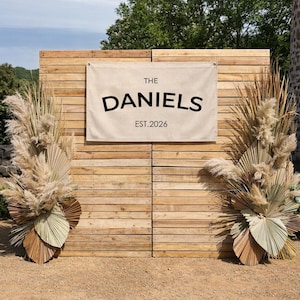 May include: A wooden backdrop with a banner that reads "THE DANIELS EST. 2026". The backdrop is flanked by dried floral arrangements. The scene is outdoors, with a natural setting.