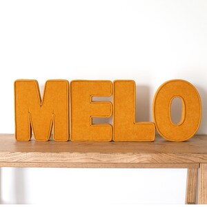 May include: Mustard yellow felt letters spelling out the name "MELO" are displayed on a wooden surface. The letters are thick and padded, with a contrasting trim. The background is a plain white wall.