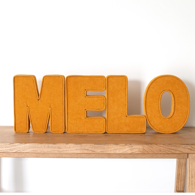 May include: Mustard yellow felt letters spelling out the name "MELO" are displayed on a wooden surface. The letters are thick and padded, with a contrasting trim. The background is a plain white wall.