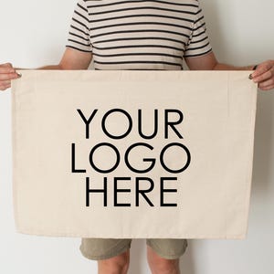 Puede incluir: Una pancarta rectangular de color crema con el texto "YOUR LOGO HERE" en letras negras grandes y en negrita. La pancarta está sostenida por una persona. La pancarta tiene ojales metálicos en cada esquina.