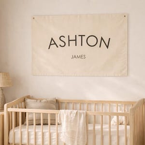 May include: A beige banner displaying the name "ASHTON" in black capital letters and the name "JAMES" below. It hangs above a wooden cot with a white blanket and pillow. A lamp and dresser are to the left.