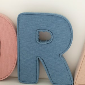May include: Close-up of a blue corduroy letter "R" pillow with a stitched border. The letter has a hole in the center. Other letters in pink and beige are partially visible.