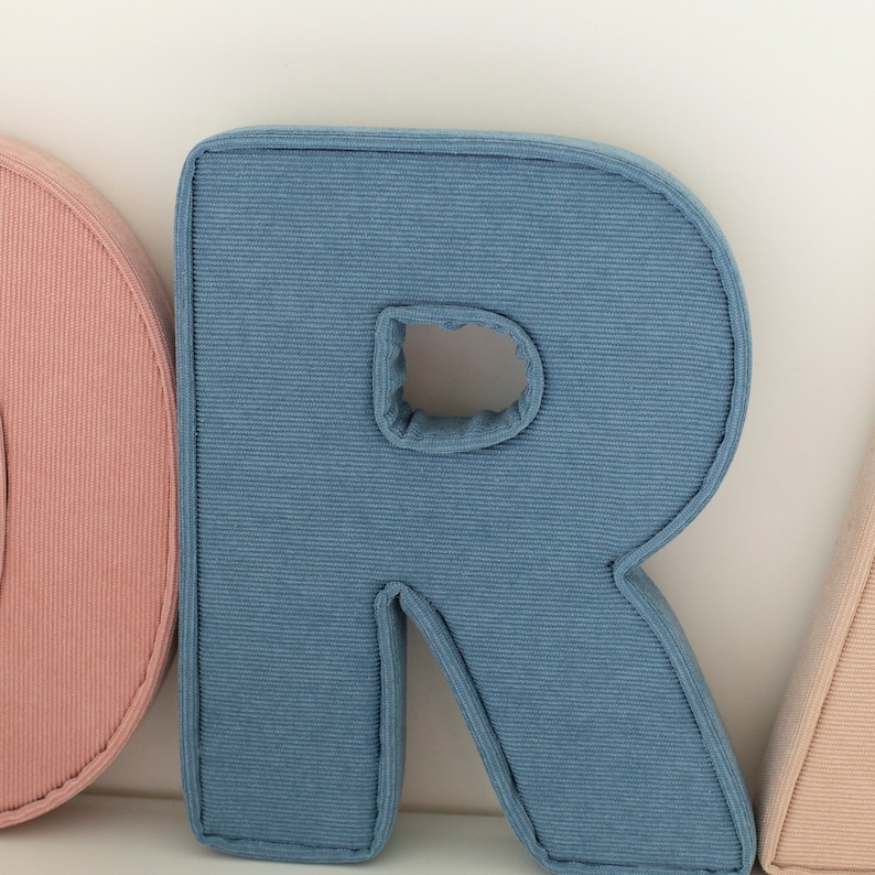 May include: Close-up of a blue corduroy letter "R" pillow with a stitched border. The letter has a hole in the center. Other letters in pink and beige are partially visible.