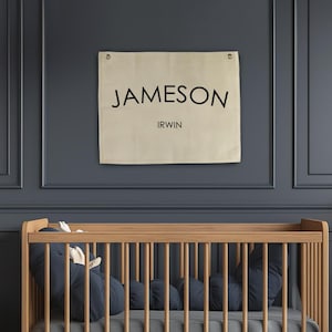 May include: A white canvas banner with the name "JAMESON IRWIN" printed in black letters. The banner is hanging on a dark grey wall above a wooden cot with a blue mattress and a blue pillow.