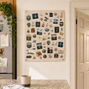 Op de afbeelding: Een rechthoekig prikbord van beige stof aan de muur, dicht bedekt met een verzameling diverse emaille pins, kleine foto's en gedrukte kaarten. Het bord is op de vier hoeken bevestigd met zilverkleurige metalen bevestigingsmiddelen.