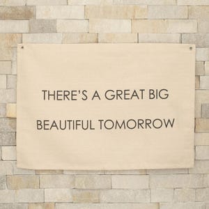 May include: A rectangular beige canvas wall banner featuring the black printed text THERE'S A GREAT BIG BEAUTIFUL TOMORROW. The banner includes two metal grommets at the top corners for hanging, providing a clean and simple display for any wall space.