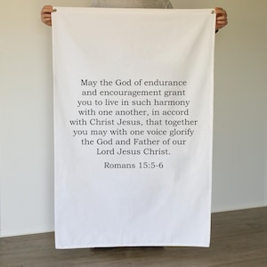 May include: White fabric banner with the text "May the God of endurance and encouragement grant you to live in such harmony...Romans 15:5-6". The banner is held up by a person.