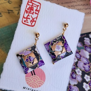 Peut inclure: Une paire de boucles d'oreilles pendantes carrées présentant un motif en papier plié avec des motifs floraux violets, noirs et dorés. Les boucles d'oreilles sont fixées sur des tiges en métal doré et présentées sur une carte blanche avec le texte Minigami Artesanía.