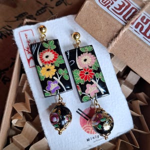 Puede incluir: Un par de pendientes rectangulares negros con diseños florales en rojo, amarillo y morado. Cada pendiente tiene un perno dorado y una cuenta redonda negra colgante con detalles dorados. Los pendientes se exhiben en una caja pequeña.