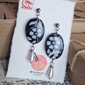 Puede incluir: Pendientes colgantes ovalados con base negra y diseño floral blanco. Cada pendiente tiene un perno plateado y un dije en forma de lágrima nacarada. Los pendientes se exhiben en una tarjeta blanca con un sello rojo y el logotipo "Minigami Artesanía".