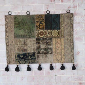 Peut inclure: Une tenture murale rectangulaire compos&eacute;e de divers segments de tapis en patchwork dans des tons de vert, bleu, marron et beige. Le textile pr&eacute;sente une s&eacute;rie de motifs g&eacute;om&eacute;triques et floraux complexes, quatre anneaux de suspension et sept pompons noirs.