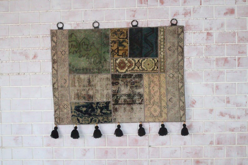 Peut inclure: Une tenture murale rectangulaire compos&eacute;e de divers segments de tapis en patchwork dans des tons de vert, bleu, marron et beige. Le textile pr&eacute;sente une s&eacute;rie de motifs g&eacute;om&eacute;triques et floraux complexes, quatre anneaux de suspension et sept pompons noirs.