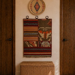 Puede incluir: Un tapiz rectangular que presenta un mosaico de tela tejida en tonos rojo, marrón, naranja y crema con patrones geométricos y flecos en la parte inferior, suspendido por tres anillos oscuros sobre un baúl de almacenamiento de mimbre.
