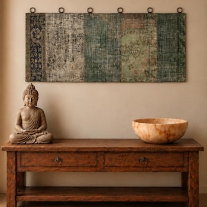 Peut inclure: Une tenture murale rectangulaire composée d'un patchwork de textiles délavés verts, bleus et sombres, suspendue par des anneaux métalliques. En dessous, une console en bois sombre à deux tiroirs porte une statue de Bouddha et un bol en pierre orange.