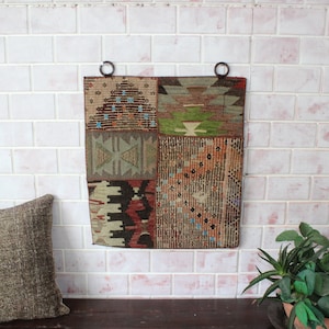 May include: A multi-colored woven tapestry with geometric patterns hangs on a white brick wall. The tapestry is made up of four squares, each with a different design. The tapestry is framed with two metal rings.