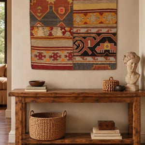 May include: A rectangular wall hanging featuring a patchwork of geometric patterns in red, orange, yellow, and dark gray. The textile is suspended by three dark metal rings. It is displayed above a wooden console table with a woven basket.