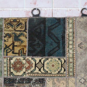 Peut inclure: Une tenture murale rectangulaire compos&eacute;e de divers fragments de tapis cousus pr&eacute;sentant des motifs g&eacute;om&eacute;triques et floraux vari&eacute;s dans des tons de vert, bleu, noir, beige et marron. Trois anneaux sombres sont fix&eacute;s au bord sup&eacute;rieur pour la suspension.