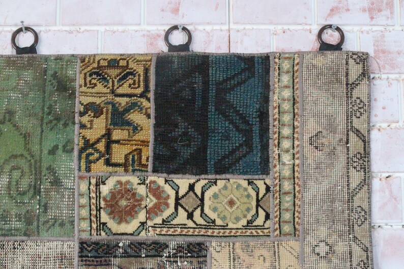 Peut inclure: Une tenture murale rectangulaire compos&eacute;e de divers fragments de tapis cousus pr&eacute;sentant des motifs g&eacute;om&eacute;triques et floraux vari&eacute;s dans des tons de vert, bleu, noir, beige et marron. Trois anneaux sombres sont fix&eacute;s au bord sup&eacute;rieur pour la suspension.