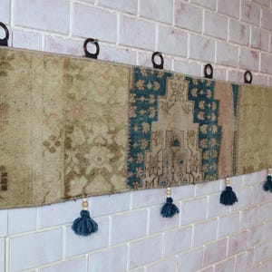 May include: A rectangular, vintage-style wall hanging with a patchwork design. The piece features sections of faded beige and teal fabric with floral and geometric patterns. Dark blue tassels hang from the bottom, and metal rings are attached at the top for hanging.