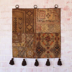 Op de afbeelding: Een vierkant wandkleed bestaande uit een patchwork van bruine, beige en oranje geweven stoffen panelen met geometrische en bloemmotieven. Het textiel hangt aan drie donkere metalen ringen en is afgewerkt met vijf donkerbruine kwastjes.