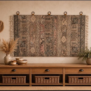 Peut inclure: Une tapisserie murale rectangulaire présentant un motif géométrique et floral aux tons sourds et multicolores avec des bords à franges, suspendue à cinq anneaux métalliques circulaires. La tapisserie affiche des nuances de brun, gris, rose et bleu.
