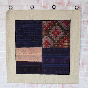 Op de afbeelding: Een vierkante wandhanger met een beige rand en een centraal patchwork-ontwerp. Het patchwork heeft secties van marineblauw, roze en bedrukte stoffen in rood, bruin en goud. De hanger wordt opgehangen door vier metalen ringen.