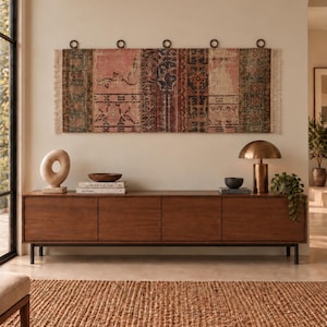 Puede incluir: Un tapiz rectangular para colgar en la pared con un diseño de patchwork en tonos rosa descolorido, rojo intenso y verde oscuro, con flecos en los laterales. El tejido cuelga de una barra horizontal mediante cinco anillas circulares oscuras.