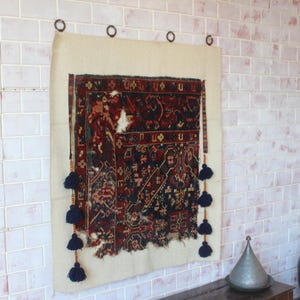 May include: A rectangular wall hanging featuring a dark red and navy blue patterned textile fragment mounted on a light beige fabric backing. The piece is suspended by four metal rings and decorated with vertical rows of dark blue yarn tassels.
