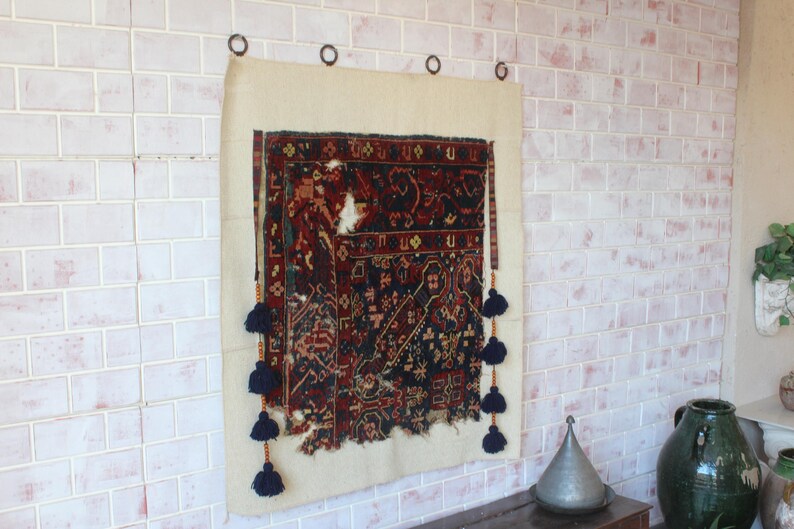 May include: A rectangular wall hanging featuring a dark red and navy blue patterned textile fragment mounted on a light beige fabric backing. The piece is suspended by four metal rings and decorated with vertical rows of dark blue yarn tassels.