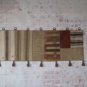 Puede incluir: Un tapiz rectangular para colgar en la pared que presenta un mosaico de paneles de tela tejida en tonos beige, marrón y rojo oscuro. El textil muestra rayas verticales, patrones geométricos y siete borlas colgantes en el borde inferior.