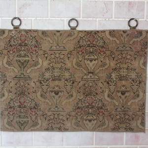 May include: A beige tapestry wall hanging with a repeating pattern of stylized floral motifs and birds. The tapestry has three metal rings at the top for hanging.